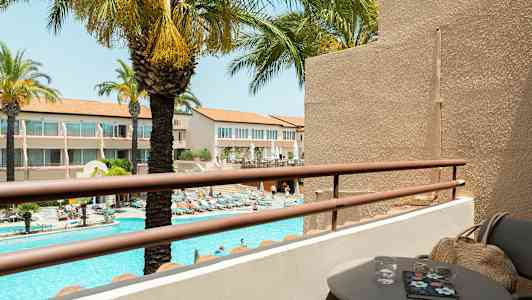 Näkymä parvekkeelta, jossa on pöytä ja tuoleja, uima-altaalle ja ympäröiviin rakennuksiin. Taustalla näkyy palmuja. (Tekoälyn luoma teksti)