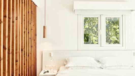 Vaalea ja ilmava makuuhuoneen sisustus, sisältäen sängyn, valkoiset vuodevaatteet ja puupaneelin sekä ikkunat, joista näkyy vehreää maisemaa. (Tekoälyn luoma teksti)