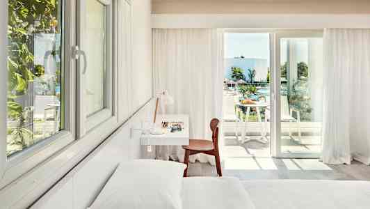 Vaalean hotellihuoneen sisätila, jossa on sänky, kirjoituspöytä ja tuoli, sekä parveke näkymällä uima-altaalle ja vihreyteen. (Tekoälyn luoma teksti)