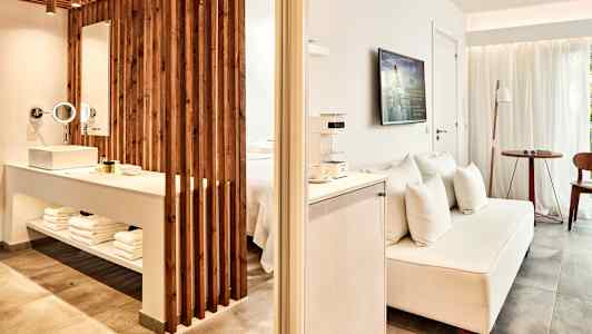 Hotellihuone avoimella suunnitelmalla, sisältäen television, sohvan ja alueen pesualtaalle ja kylpyhuonetarpeille. (Tekoälyn luoma teksti)