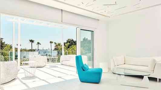 Moderni olohuone, jossa on suuret ikkunat ja näkymä aurinkoiselle terassille, jota ympäröivät palmut. (Tekoälyn luoma teksti)