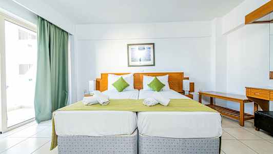 Kaksi yhden hengen sänkyä vihreällä päiväpeitteellä ja valkoisilla tyynyillä, vaaleasti sisustetussa hotellihuoneessa. (Tekoälyn luoma teksti)