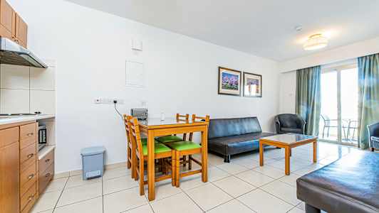 Olohuone, jossa on huonekaluja, kuten pöytä tuoleineen, sohva ja nojatuoli. Keittiö näkyy nurkassa ja parveke ovi taustalla. (Tekoälyn luoma teksti)