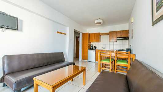 Sisätilan näkymä, jossa on kaksi ruskeaa sohvaa, lasipöytä ja puukaapeilla varustettu keittiö ja ruokailualue. (Tekoälyn luoma teksti)