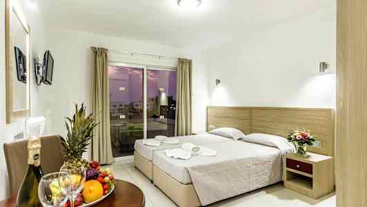 Kuva hotellihuoneesta, jossa on kaksi erillistä vuodetta, pöytä hedelmillä ja juomalla sekä parveke näkymällä. (Tekoälyn luoma teksti)