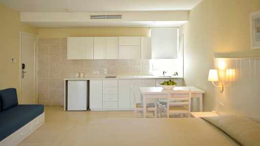 Valoisa ja moderni studio, jossa keittiöalue, valkoisia kaappeja, pieni pöytä tuoleineen ja istumakaluste beigellä seinällä. (Tekoälyn luoma teksti)