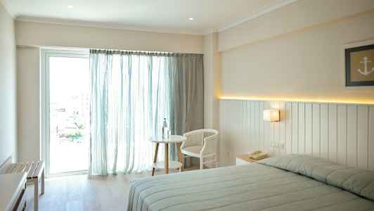 Kuva vaaleasta hotellihuoneesta, jossa on sänky, pöytä ja tuoli lähellä ikkunaa, jossa on vaaleat verhot. (Tekoälyn luoma teksti)