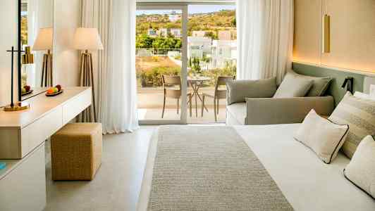 Kuva hotellihuoneesta, jossa on sänky, sohva ja kirjoituspöytä, sekä parveke näkymällä asuinalueelle. (Tekoälyn luoma teksti)