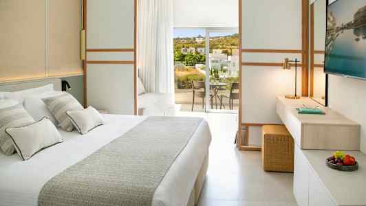 Valoisa hotellihuone, jossa on suuri sänky, työtila ja parveke näkymällä vehreään maisemaan ja rakennuksiin. (Tekoälyn luoma teksti)