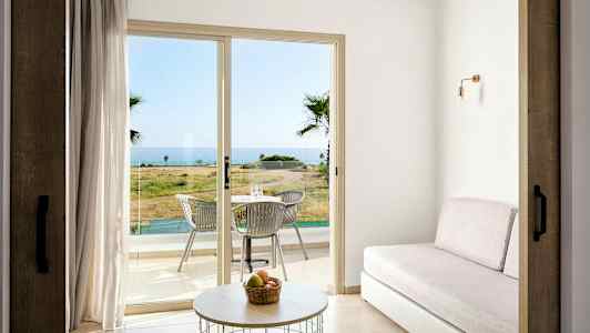 Huone, jossa on sohva, pöytä ja lasiovet, jotka johtavat terassille, jossa on pöytä ja tuolit, meri näköalalla. (Tekoälyn luoma teksti)