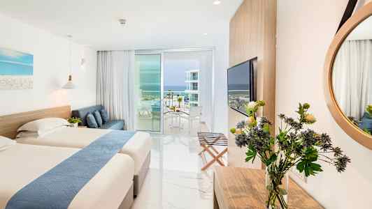 Hotellihuone, jossa on kaksi sänkyä, sohva, pöytä kukilla, iso peili ja parveke, josta on näkymä merelle. (Tekoälyn luoma teksti)