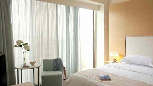 Makuuhuone, jossa on pedattu sänky, tuoli, pöytälamppu ja ikkuna, jossa vaaleat verhot. (Tekoälyn luoma teksti)