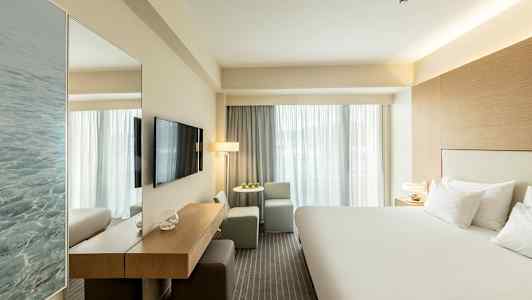 Hotellihuone, jossa iso sänky, TV seinällä ja istuinryhmä ikkunan vieressä. (Tekoälyn luoma teksti)