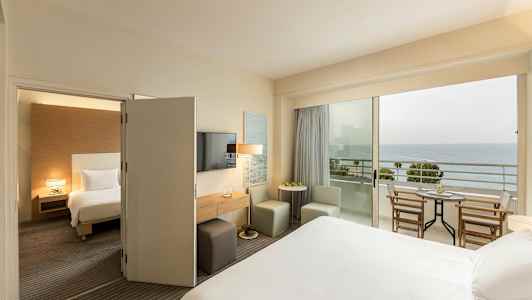 Hotellihuone, jossa on sänky, istuimia ja pöytä, sekä parveke merinäköalalla. (Tekoälyn luoma teksti)