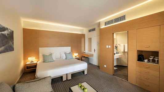 Moderni hotellihuone, jossa on parivuode, istuinryhmä ja kirjoituspöytä. Oikealla on vaatekaappi ja ovi, joka johtaa kylpyhuoneeseen. (Tekoälyn luoma teksti)