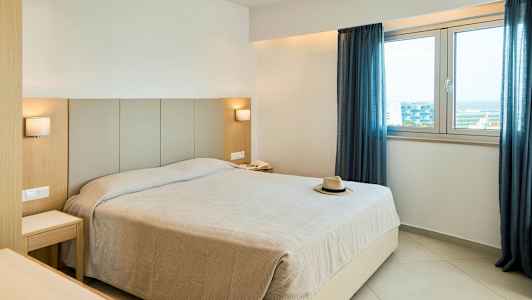 Hotellihuone, jossa suuri sänky, kaksi yöpöytää, kaksi lamppua, ikkuna sinisin verhoin ja hattu sängyllä. (Tekoälyn luoma teksti)