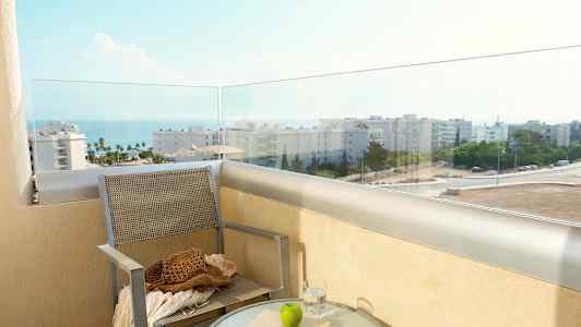 Parveke, jolta on näkymä merelle ja rakennuksiin, tuoli ja pöytä vedellä, kirjalla ja omenalla. (Tekoälyn luoma teksti)