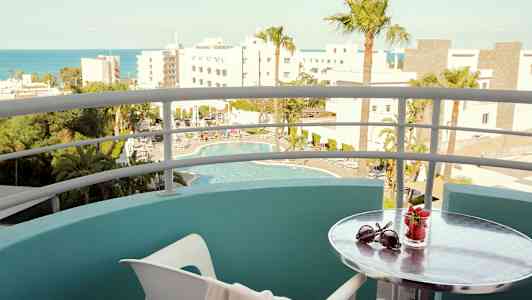 Näkymä parvekkeelta uima-altaalle ja ympäröiviin rakennuksiin; pienellä pöydällä on aurinkolasit ja mansikoita sisältävä lasi. (Tekoälyn luoma teksti)