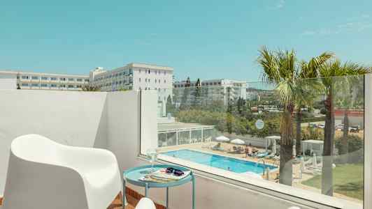 Parveke, jossa valkoisia tuoleja ja pöytä, näkymä uima-altaalle ja ympäröiviin rakennuksiin. (Tekoälyn luoma teksti)