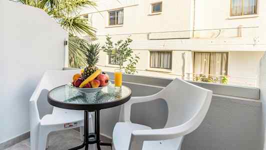 Parveke, jossa pöydällä on hedelmäkulho ja lasillinen mehua, ympärillä kaksi valkoista tuolia. (Tekoälyn luoma teksti)