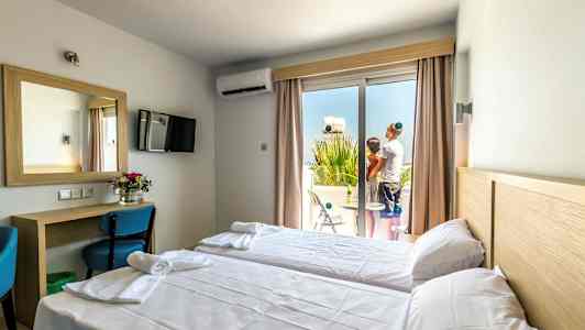 Hotellihuone, jossa on kaksi yhden hengen vuodetta, kirjoituspöytä ja peili, sekä parveke, jossa seisoo kaksi henkilöä. (Tekoälyn luoma teksti)