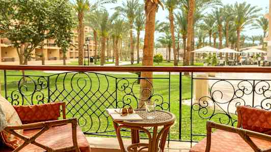 Parveke, jossa on tuoleja, pöytä lasin ja aurinkolasien kanssa, näkymä puutarhaan palmupuiden keskellä. (Tekoälyn luoma teksti)