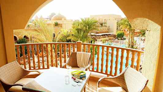 Parveke, jossa kalusteita ja näkymä palmuihin, uima-altaaseen ja hiekanvärisiin rakennuksiin. (Tekoälyn luoma teksti)