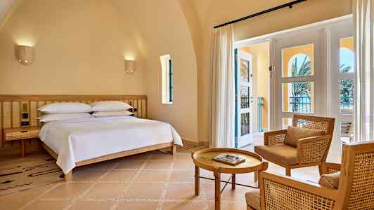 Huone, jossa on sänky, kaksi tuolia ja pöytä sekä lasiovi, joka johtaa parvekkeelle. (Tekoälyn luoma teksti)