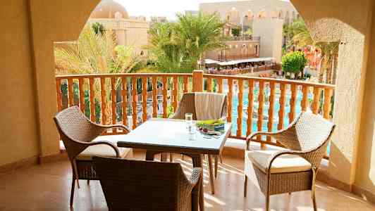 Näkymä parvekkeelta, missä on pöytä ja tuolit, ja näkymä altaalle sekä palmuja arkkitehtonisen rakennuksen luona. (Tekoälyn luoma teksti)