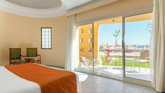 Huone, jossa on sänky ja oranssinväriset vuodevaatteet, kaksi tuolia ja pöytä sekä näkymä terassille, jossa on vihreää ja aurinkotuoleja. (Tekoälyn luoma teksti)