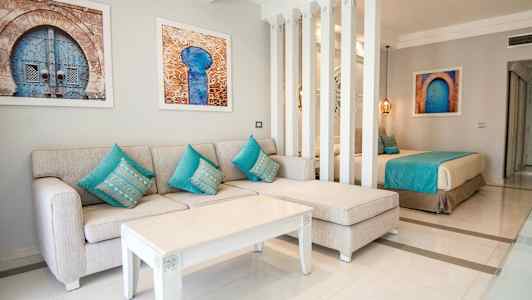 Oleskelutila, jossa beige sohva ja turkoosit tyynyt, valkoinen sohvapöytä ja taideteoksia seinällä, osittain pylväillä erotettu sänkyalueesta. (Tekoälyn luoma teksti)