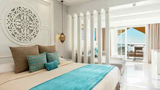 Hotellihuone, jossa on vaaleat seinät, koristeltu sänky sinisillä ja beigeillä vuodevaatteilla sekä parveke merinäköalalla. (Tekoälyn luoma teksti)