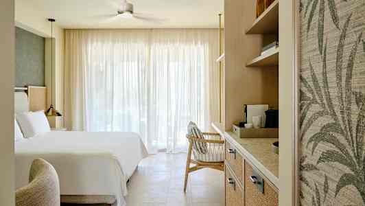 Hotellihuone, jossa on iso sänky, nojatuoli ja hyllyjä kahvinkeittimellä, valaistuna luonnonvalolla suurten verhojen läpi. (Tekoälyn luoma teksti)