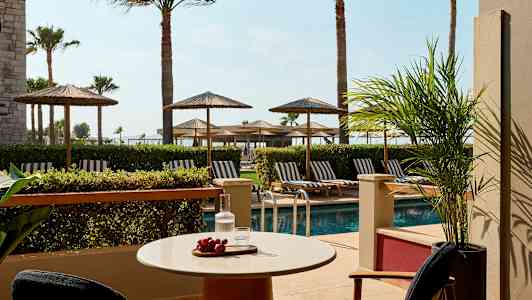 Terassi, jossa pöytä ja tuolit, näkymä uima-altaalle ja aurinkotuoleihin varjojen alla. (Tekoälyn luoma teksti)