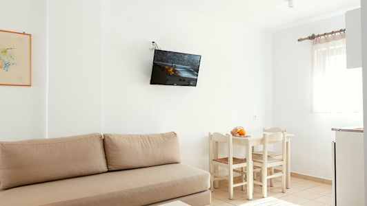 Vaalea huone, jossa beige sohva, seinään kiinnitetty TV, valkoinen ruokailuryhmä tuoleineen ja ikkuna vaaleilla verhoilla. (Tekoälyn luoma teksti)