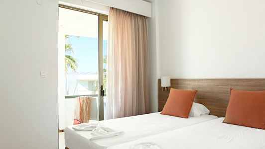 Hotellihuone, jossa on kaksi yhden hengen vuodetta, oransseja tyynyjä ja lasiovi parvekkeelle. (Tekoälyn luoma teksti)