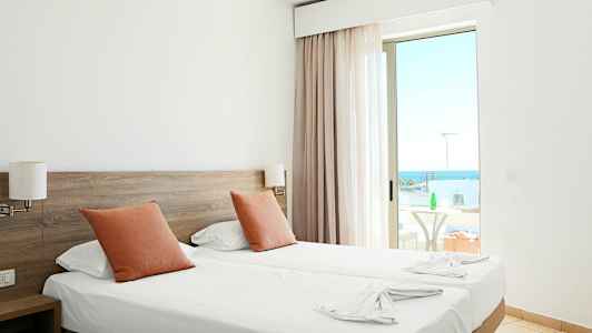 Hotellihuone, jossa on kaksi erillistä sänkyä, oranssit tyynyt ja parveke merinäköalalla. (Tekoälyn luoma teksti)