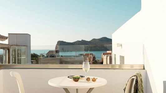 Näkymä terassilta, pöytä, tuoli, viinilasi ja välipalaa, taustalla meri ja vuoria. (Tekoälyn luoma teksti)