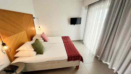 Kuva hotellihuoneesta, jossa on parisänky, puupaneeli sängynpäätynä, punaisia ja vihreitä koristetyynyjä, riippuvalaisimia, pieni pöytäpuhelin ja verhot. Televisio on seinällä. (Tekoälyn luoma teksti)