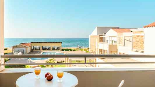 Näköala parvekkeelta, jossa on pöytä ja kaksi lasia sekä kaksi omenaa, kohti uima-allasta, rakennuksia ja merta. (Tekoälyn luoma teksti)