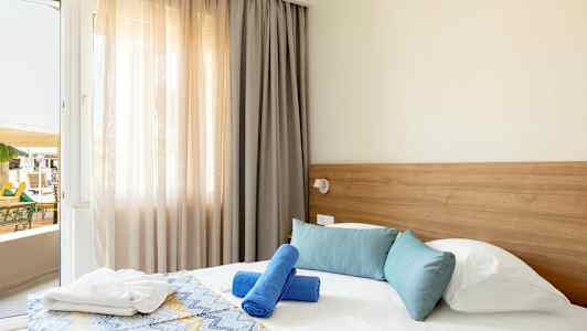 Hotellihuone, jossa on sänky, sinisiä ja valkoisia tyynyjä sekä kaksi sinistä kylpypyyhettä. Ikkunapariovi on auki parvekkeelle. (Tekoälyn luoma teksti)