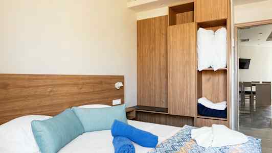 Hotellihuone, jossa sängyssä on sinisiä tyynyjä, sinisiä ja valkoisia pyyhkeitä sekä värikäs kuviollinen päiväpeitto. Taustalla näkyy puunvärinen kaappi ja ovi toiseen huoneeseen. (Tekoälyn luoma teksti)