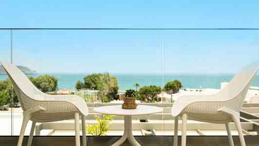 Näkymä parvekkeelta, jossa on kaksi valkoista tuolia ja pöytä pienellä kasvilla, sekä merimaisema ja vehreä tausta. (Tekoälyn luoma teksti)
