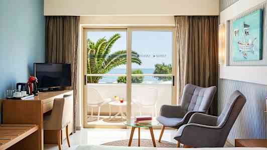 Hotellihuone, jossa istuma-alue ja näkymä parvekkeelle, jossa pöytä ja tuolit sekä merinäköala. (Tekoälyn luoma teksti)