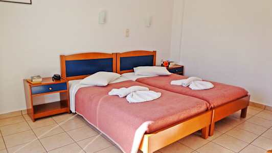 Kaksi erillistä vuodetta taiteltujen pyyhkeiden ja vaaleanpunaisten peitteiden kanssa, kaksi yöpöytää ja laattalattia hotellihuoneessa. (Tekoälyn luoma teksti)