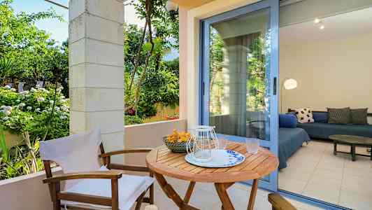 Ulkoiluterassi, jossa on puupöytä ja tuoleja, ympäröi vihreät kasvit. Lasinen ovi johtaa huoneeseen, jossa on istuimet. (Tekoälyn luoma teksti)