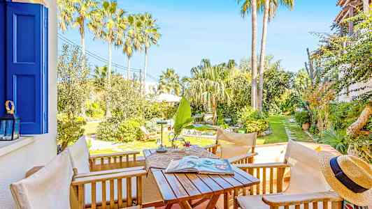 Terassi, jossa puutarhakalusteita, näkymä aurinkoiseen puutarhaan, jossa palmuja ja kasvillisuutta. (Tekoälyn luoma teksti)