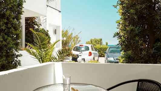 Terassi, jolla pöytä, kirja ja lasi. Ulkopuolella pysäköityjä autoja ja kasvillisuutta. (Tekoälyn luoma teksti)