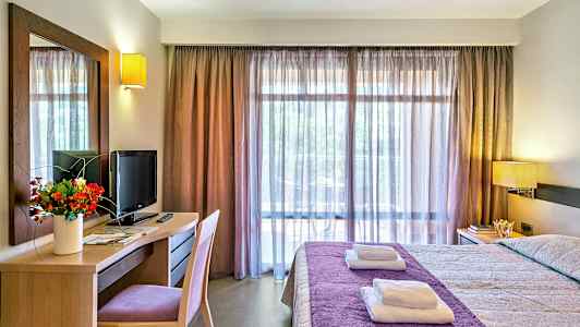 Hotellihuone, jossa sängyllä on violetti päiväpeite, työpöydällä televisio ja kukkakimppu, sekä suuret ikkunat, joissa on läpikuultavat verhot. (Tekoälyn luoma teksti)