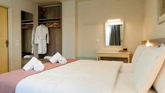 Hotellihuone, jossa on parivuode, valkoiset vuodevaatteet ja vaaleanpunainen huopa. Taustalla näkyy vaatekaappi, jossa roikkuu vaatteita, sekä peili. (Tekoälyn luoma teksti)